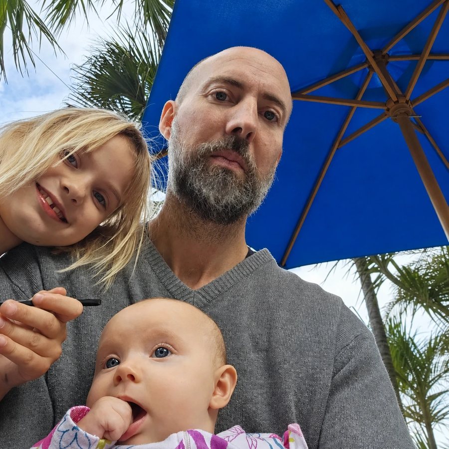 Nicholas Harvey, founder of Salty Air Home Cleaning, outdoors with his two young children under a blue umbrella, palm trees behind them.